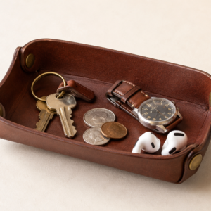 Brown leather catchall tray with brass hardware holding keys coins a watch and earbuds on a clean light background